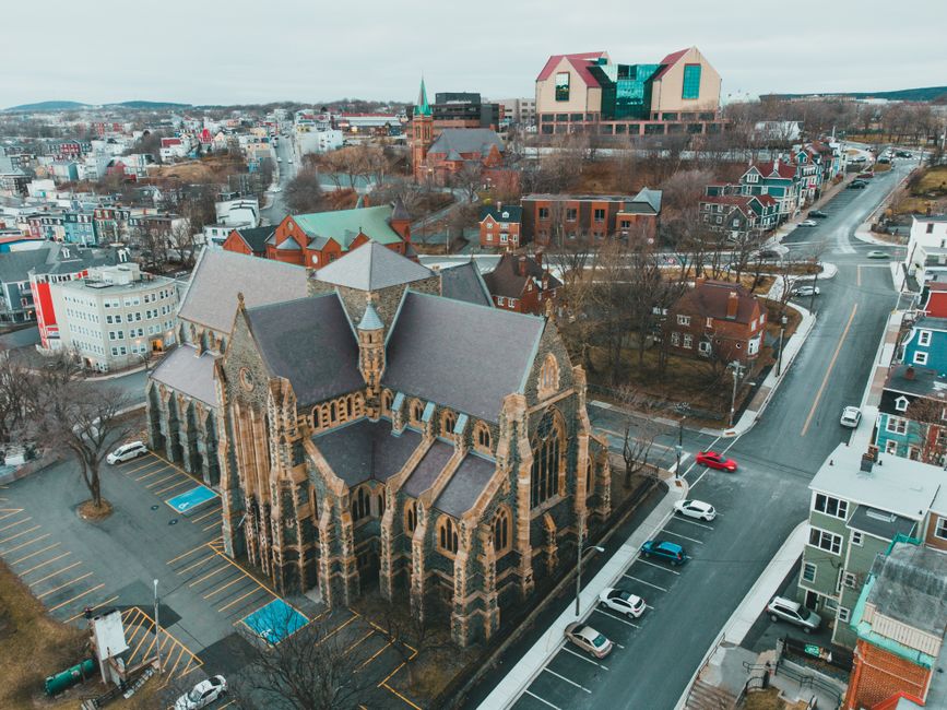An aerial view of St Johns Church, a historic stone building with pointed arches, stained glass windows, and a steeply pitched roof, situated among a mix of residential houses and modern commercial st