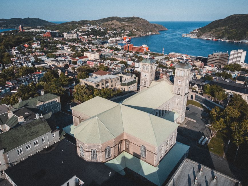 An aerial view of St Johns Church, a historic stone building with pointed arches, stained glass windows, and a steeply pitched roof, situated among a mix of residential houses and modern commercial st